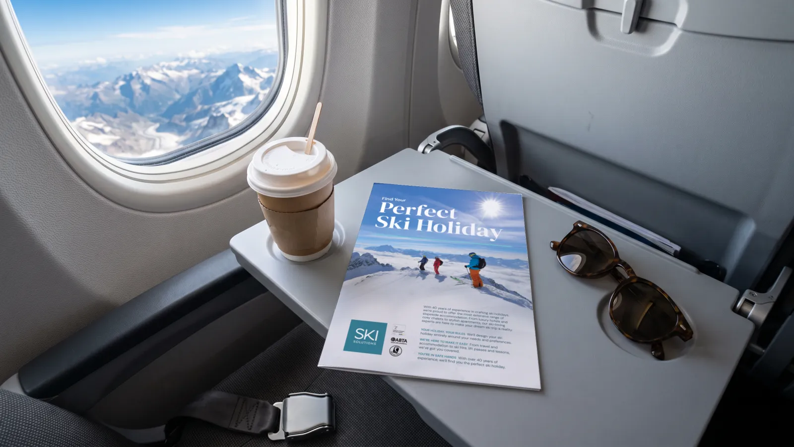 The Ski Solutions Find Your Perfect Ski Holiday advert on an aeroplane tray table by the window over the Alps, in a portfolio visualisation.