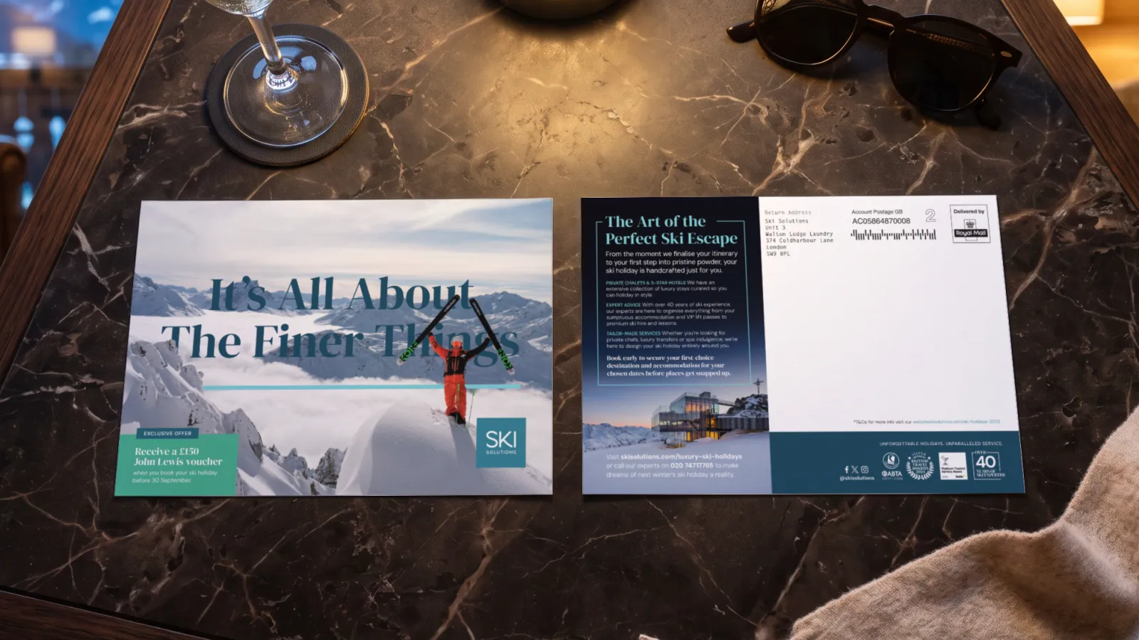 The Ski Solutions It’s All About The Finer Things luxury mailer, front and back, on a marble table in an alpine hotel suite beside a glass of champagne, in a portfolio visualisation.