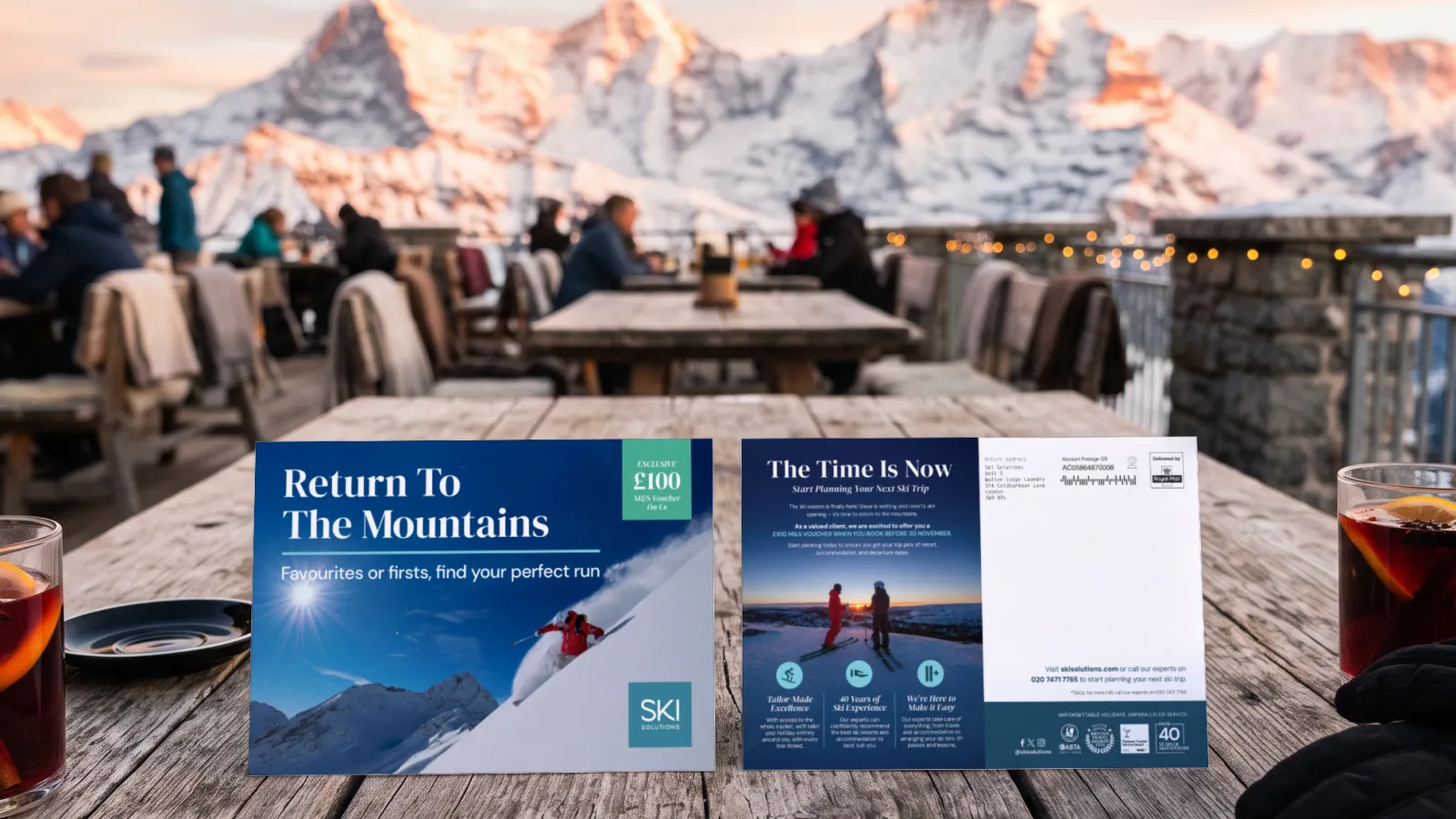 The Ski Solutions November mailer, front and back, standing on a mountain terrace table at sunset between two glasses of mulled wine, in a portfolio visualisation.