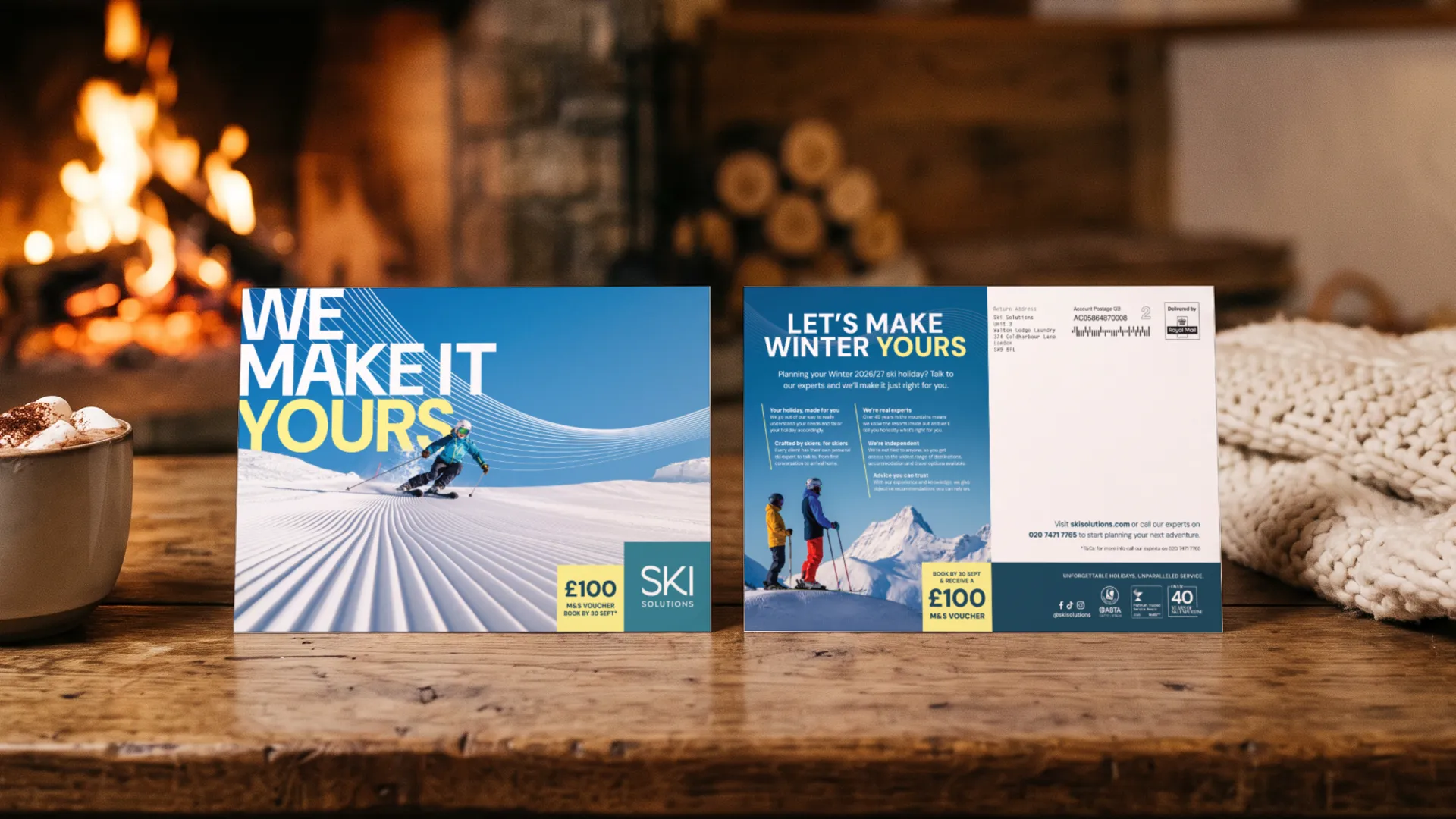 The Ski Solutions We Make It Yours direct mail, front and back, standing on a chalet coffee table in front of a log fire, with a mug of hot chocolate, in a portfolio visualisation.